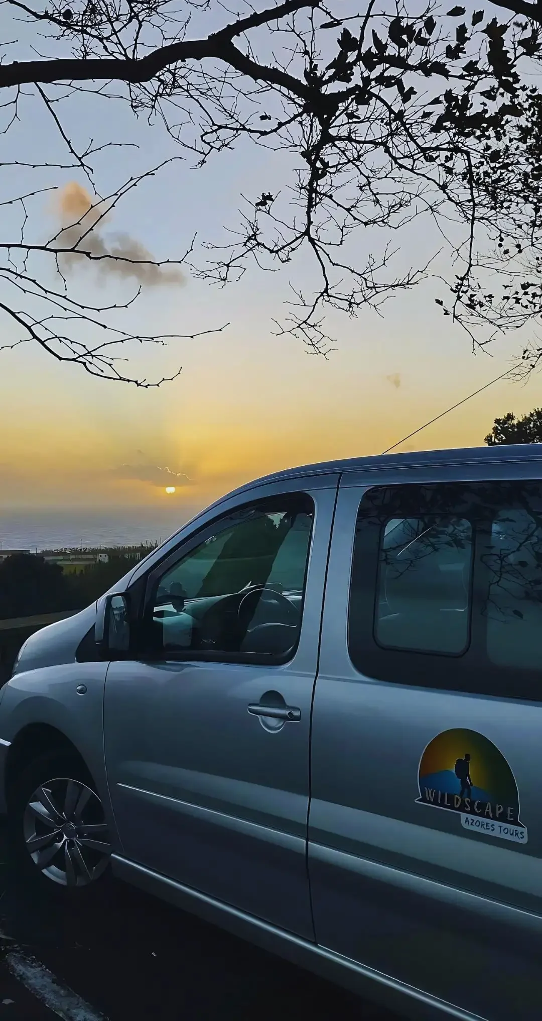 The view of the left face of a van with the sunset over the sea on the horizon.