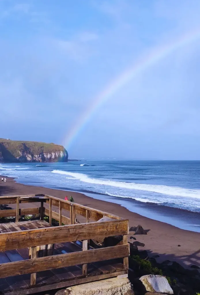 A dark sand beach with waves breaking and a rainbow on the background.