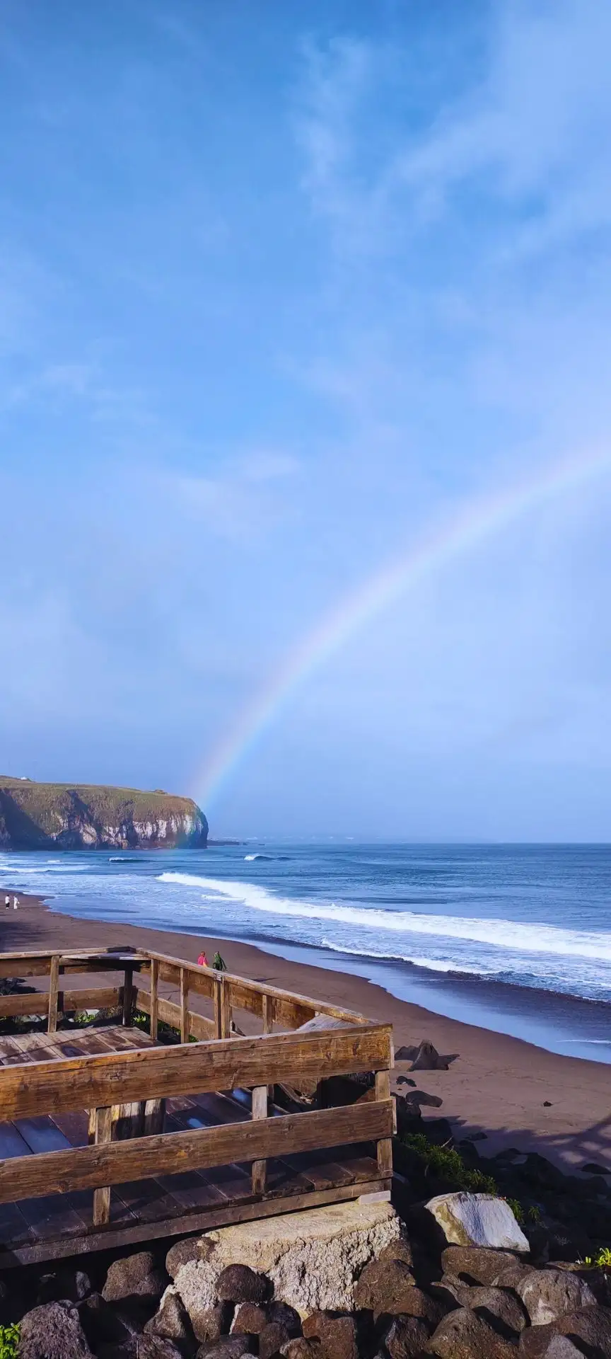 A dark sand beach with waves breaking and a rainbow on the background.