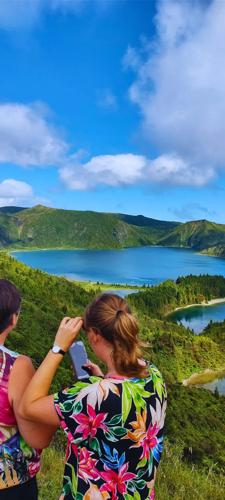 Two women take photos of a big a lagoon surrounded by dormant volcano walls.