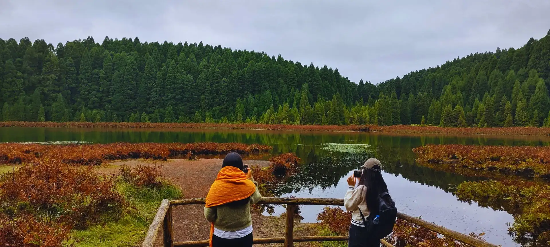 Two girls photographing a green lagoon surrounded by trees.