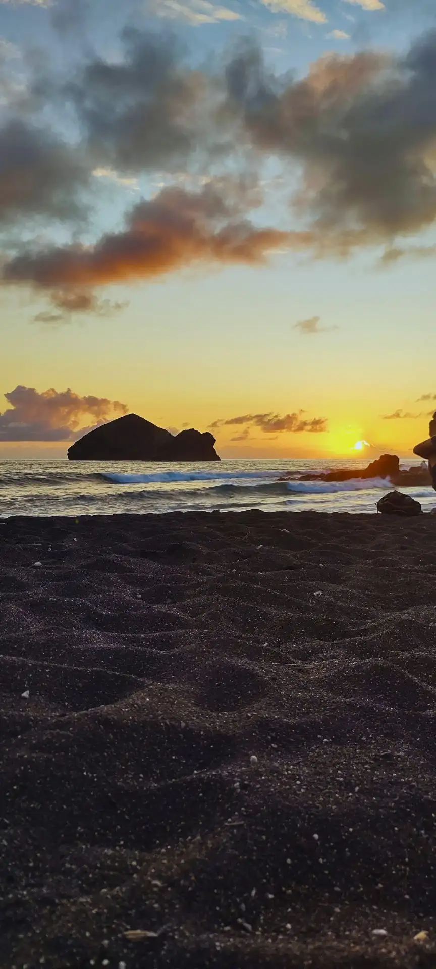 A dark sand beach with the ocean, islets and a sunset on the background.