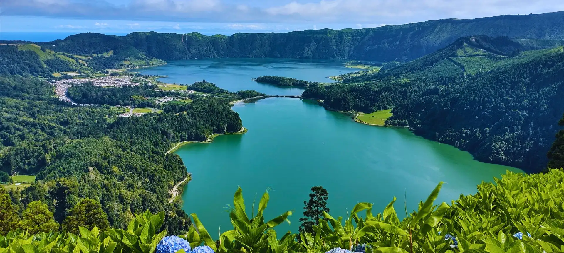 An high view to a big green and blue lagoon, divided in the middle by a bridge and surrounded by collapsed walls of a dormant volcano.