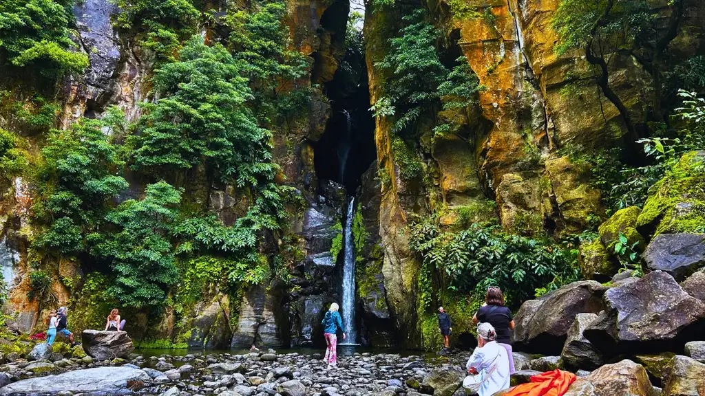 A waterfall is seen among tall walls of rock, surrounding vegetation is lush. A group of people contemplate the views and take photos.