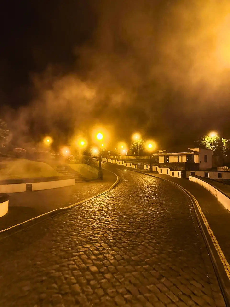 A cobblestone street of Furnas at night, above the lamp lights reflect on the floating steam.