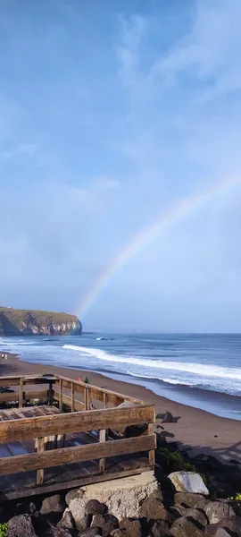 A dark sand beach with waves breaking and a rainbow on the background.